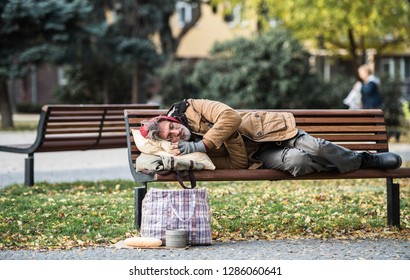 Homeless beggar man with a bag lying on bench outdoors in city, sleeping.