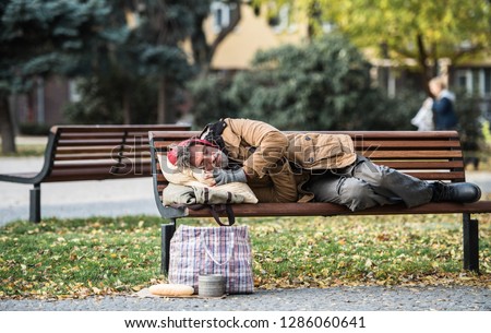 Homeless beggar man with a bag lying on bench outdoors in city, sleeping.