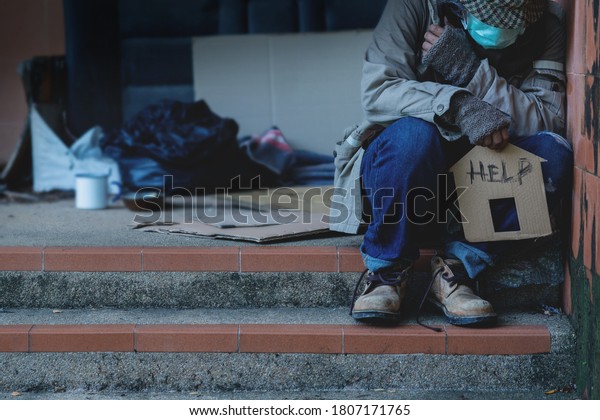 Homeless begging man with a medical mask sits\
on the steps holding home shape cardboard, The word \
