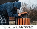 homeless elderly old Caucasian man rummages for food and garbage in a trash can in a park in autumn