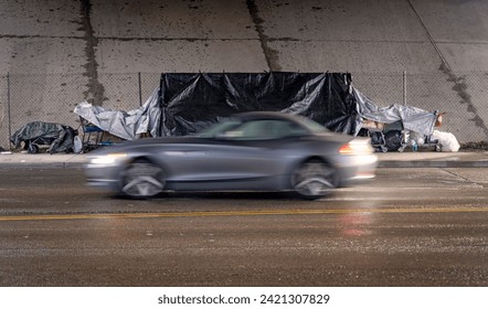 A homeless encampment under a bridge with an expensive car blurred in the foreground on a rain soaked street