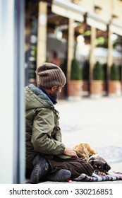 A homeless guy is sitting in the ground with a dog in his feet. He is begging for money in the streets. The man is unidentified. The image has a vintage effect applied.