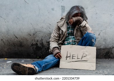 Homeless, Long-haired Asian Man Sits Hopelessly Leaning Against A Wall As There Is No One To Help Him With Work And Food In His Hand Holding A Sign For Help. Homeless Sleep On Streets