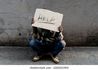 A Homeless, Long-haired Asian Man Sits Hopelessly Leaning Against A Wall As There Is No One To Help Him With Work And Food In His Hand Holding A Sign For Help. Homeless Sleep On Streets