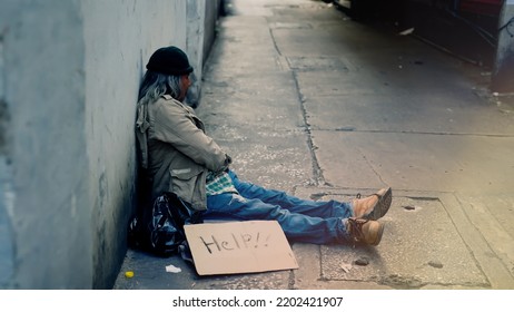 A Homeless, Long-haired Asian Man Sits Hopelessly Leaning Against A Wall As There Is No One To Help Him With Work And Food In His Hand Holding A Sign For Help. Homeless Sleep On Streets