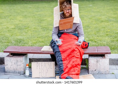 Homeless Man With Box On His Head Sits On The Bench Wrapped With Sleeping Bag Begging Money In City Park, Outdoor. Cardboard With Text 