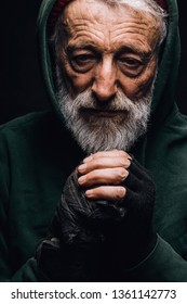 Homeless man dressed in green warm old big-sized clothing with hood on shivers from cold, tries to warm up, looking down with mournful expression