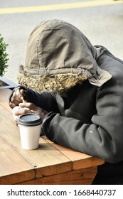 Homeless Man Eating At Outdoor Table With Winter Hooded Coat