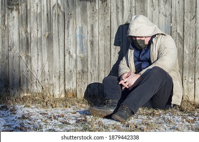 Homeless. A Man In Ragged Clothes And A Medical Mask Sits By A Wooden Fence. COVID-19. Close Up.