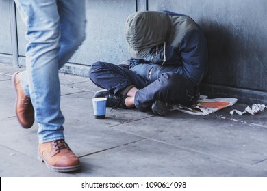 Homeless Man Sitting On The Street In The Shadow Of The Building And Begging For Help And Money. Problems Of Big Modern Cities.