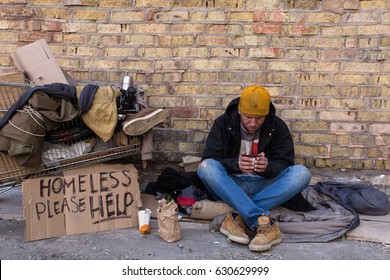Homeless man sitting on the street, near the wall and cart with his stuff. Holding cup of coffee and looking in it.