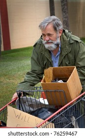 Homeless man wearing an old army coat pushes a shopping cart holding his possessions.