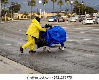 A homeless person in a yellow plastic jacket pushing a shopping cart in the rain 