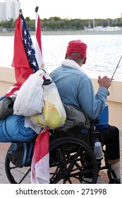 A homeless veteran in a wheelchair, with an american flag,  staring over the side of a pier at the water.