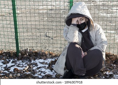 Homeless. A Woman In Dirty Ragged Clothes And A Medical Mask Is Sitting On The Ground. Close Up.