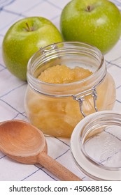 Homemade Apple Sauce In Clear Glass Jar With A Sealed Lid, Wood Spoon  And Two Green Apples On A Blue-white Table Cloth