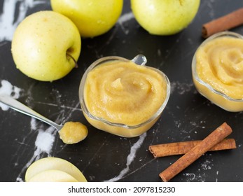 Homemade applesauce (apple puree or mousse) in small glass bowls with organic golden delicious apples and cinnamon sticks on a black textured table. Creamy and healthy baby food.