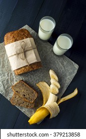 Homemade Banana Bread With Slices, Two Glasses Of Milk And Banana On Baking Paper On Dark Background. Overhead View.