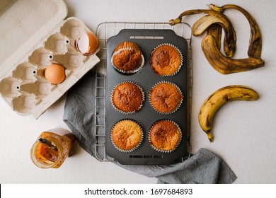 Homemade Banana And Honey Muffins In Mufin Tray Cooling On Rack, Eggs, Banana Peel, Honey On Kitchen Tabletop. Flat Layout, Horizontal Orientation