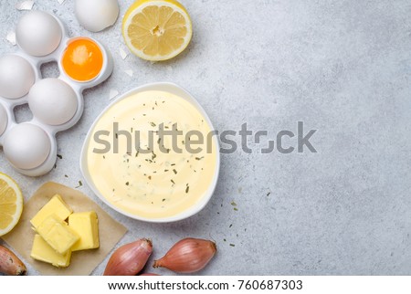 Homemade basic french sauce bearnaise in a white bowl with ingredients, butter, shallot, lemon, eggs, on a light blue stone background with copy space, top view