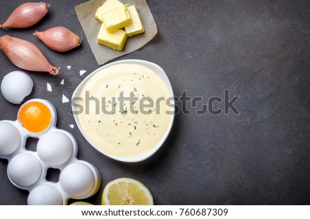 Homemade basic french sauce bearnaise in a white bowl with ingredients, butter, shallot, lemon, eggs, on a black slate stone background with copy space, top view
