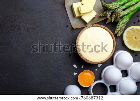 Homemade basic french sauce hollandaise in a wooden bowl with ingredients, butter, asparagus, lemon, eggs, on a black slate stone background with copy space, top view