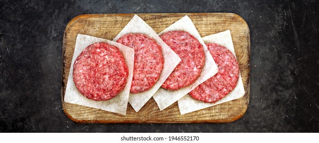 Homemade Beef Burgers On Wooden Board, Top View. Four Raw Ground Beef Meat Steak Burgers Cutlets On Black Table Background, Overhead View. Farmers Uncooked Beef Burgers For BBQ Grilling Or Frying.