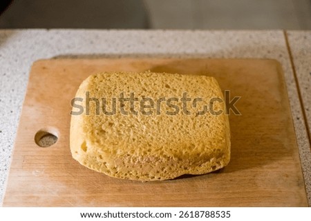 Homemade bread rests upside down upon wooden board, placed over kitchen counter near hole designed for grip, showing porous texture, golden crust, baked edges, smooth surface.