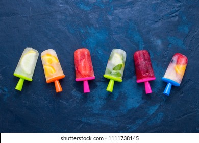Homemade bright Fruity popsicle with strawberry, cherry, lemon, orange, lemon and mint flavor on a dark blue background. Flat lay, top view.