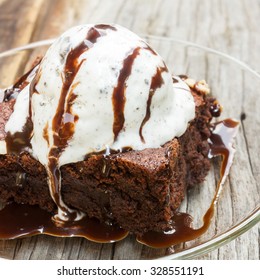 Homemade Brownies With Ice Cream On Top And Chocolate Sauce Are Topping. On Glass Plate Over Wood Table.