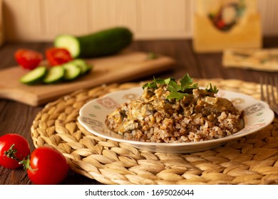 Homemade Buckwheat Porridge With Mushrooms In Sour Cream, With Parsley And Vegetables, Cucumber, Cherry Tomato On White Plate On Natural Serving Tray On Brown Wooden Table In Cozy Kitchen