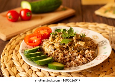 Homemade Buckwheat Porridge With Mushrooms In Sour Cream, With Parsley And Vegetables, Cucumber, Cherry Tomato On White Plate On Natural Serving Tray On Brown Wooden Table In Cozy Kitchen