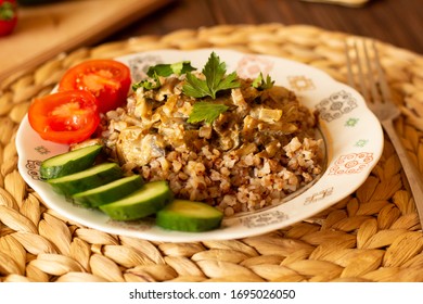 Homemade Buckwheat Porridge With Mushrooms In Sour Cream, With Parsley And Vegetables, Cucumber, Cherry Tomato On White Plate On Natural Serving Tray On Brown Wooden Table In Cozy Kitchen