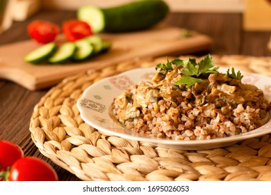 Homemade Buckwheat Porridge With Mushrooms In Sour Cream, With Parsley And Vegetables, Cucumber, Cherry Tomato On White Plate On Natural Serving Tray On Brown Wooden Table In Cozy Kitchen