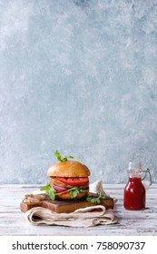 Homemade Burger In Classic Bun With Tomato Sauce, Arugula, Meat, Cheese, Onion, Bottle Of Ketchup On Wood Serving Board Over White Wooden Plank Table. Rustic Style. Homemade Fast Food.