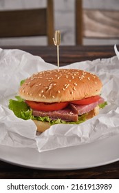 Homemade Burger Stuffed With Dried Red Fish, Tomato, Onion, Pickled Cucumber, Lettuce Leaves In Paper On A Plate Close-up. Fast Food. Vertical Orientation, No People