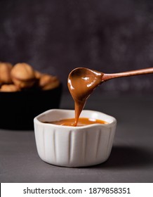 Homemade Caramel Sauce Flows From A Wood Spoon Into A White Bowl On Dark Background. Front View And Close Up