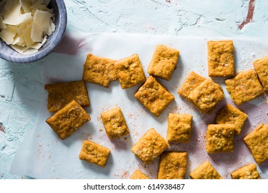 Homemade Cheesy Turmeric Crackers On A Textured Blue Plaster Surface With Copper Accents And Small Bowl Of Shaved Parmesan Cheese.