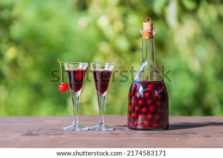 Homemade cherry brandy in two glasses and in a glass bottle on a wooden table in a summer garden, close up