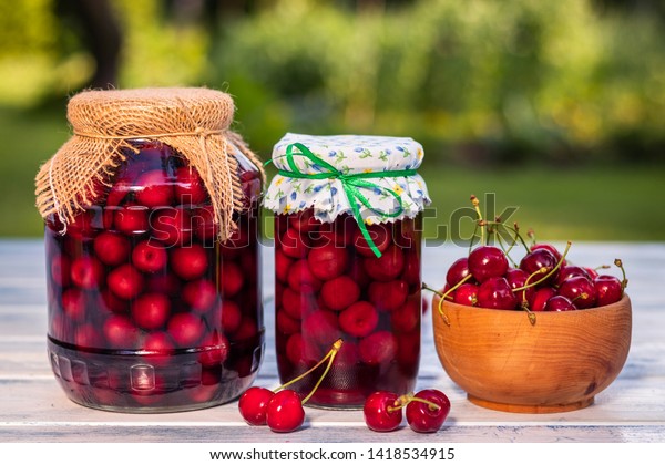 Homemade cherry fruit compote in glass jar and fresh\
harvested red cherries in wooden bowl. Preserved organic food from\
garden. 