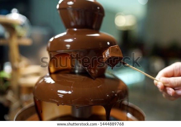 Homemade chocolate fountain fondue with\
marshmallow on a skewer dripping in chocolate sauce on blurred\
background and copy\
space.