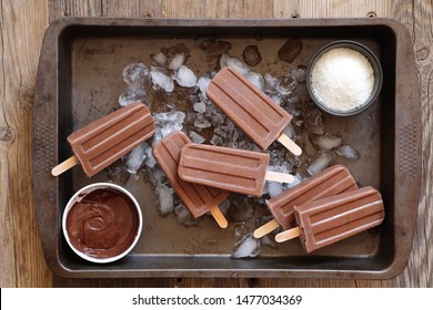 Homemade chocolate popsicles on ice in rustic tray on wooden background. Chocolate fudgesicles with chocolate and coconut flakes - horizontal image 