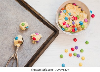 Homemade Cookie Dough With Colorful Chocolate Candies In Bowl, Ice Cream Scooper And Scoops Of Cookie Dough On Baking Sheet On White Wooden Background