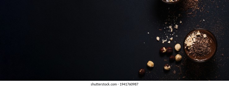 Homemade dark chocolate mousse with hazelnuts in glasses on dark grey table, close up, top view, selective focus, banner