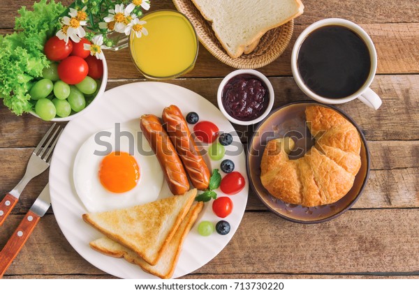 Homemade delicious American breakfast with sunny side up fried egg, toast, sausage, fruits, vegetable, strawberry jam, croissant, black coffee and fresh orange juice in top view flat lay style.
