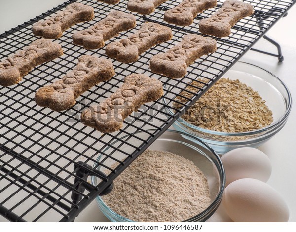 Homemade dog treats with the word Woof on them cool on a rack, accompanied with ingredients eggs, oats, and whole wheat flour