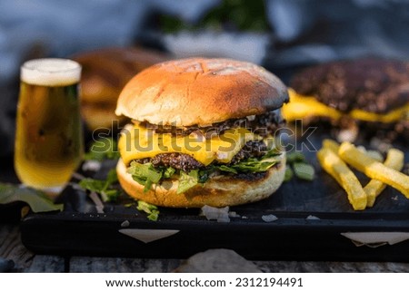 
homemade double smash burger with cheese, lettuce, onion and dressing on a cutting board. And a pile of fries on one side and a glass of cold beer on the other