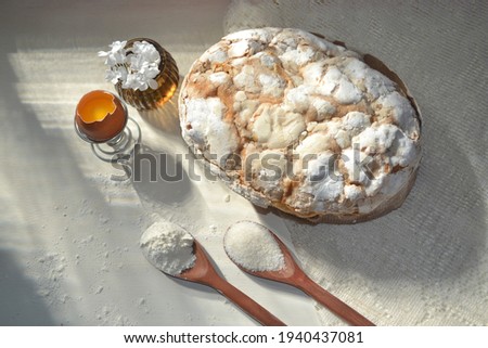 Homemade Easter cake (Colomba Pasquale or Colomba Pascal) on a rustic white wooden table. Lace tablecloth, eggs,flowers, wooden spoon with sugar and flour in the composition. Happy easter concept.