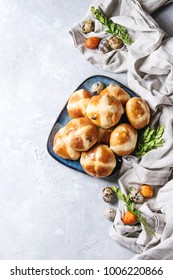 Homemade Easter traditional hot cross buns on blue plate with textile and colored quail eggs over white texture background. Top view, copy space