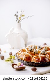 Homemade Easter traditional hot cross buns in ceramic dish with blossom flowers, rabbit, chocolate candy and quail eggs on white marble table. Easter holiday baking and decor